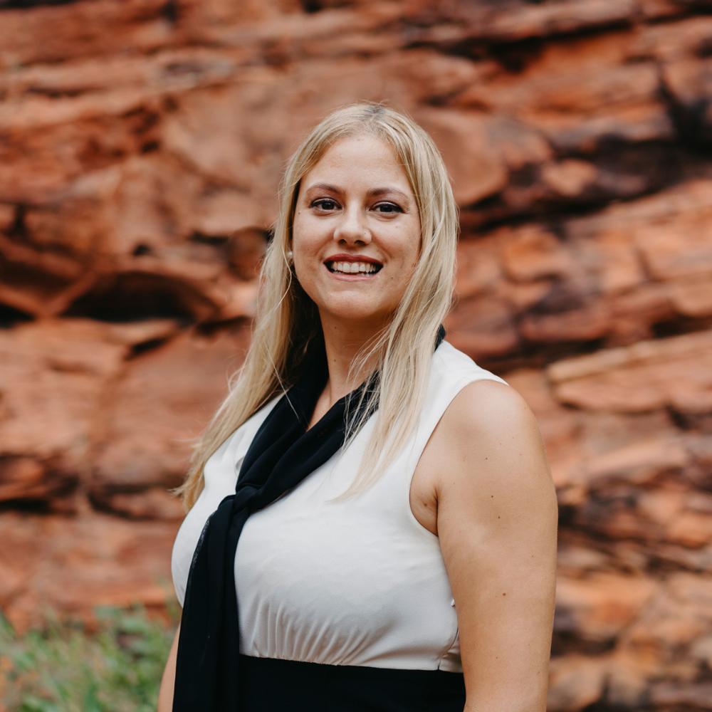 Portrait of Anita Sutherland, a smiling woman with blonde hair, wearing a white sleeveless top and black scarf, with a red rocky backdrop indicative of the Kimberley landscape.