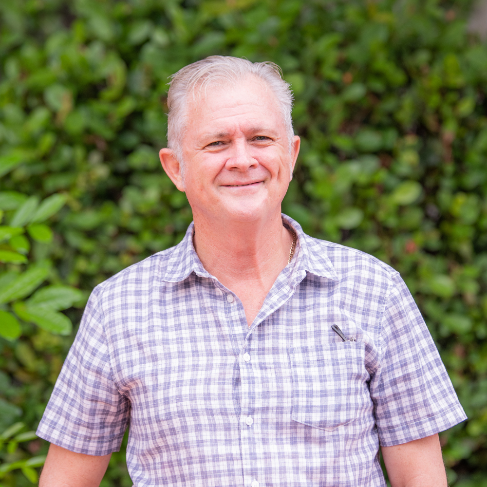 Chris Maher, a man with short grey hair, wearing a checkered shirt, smiling in front of a lush green hedge.